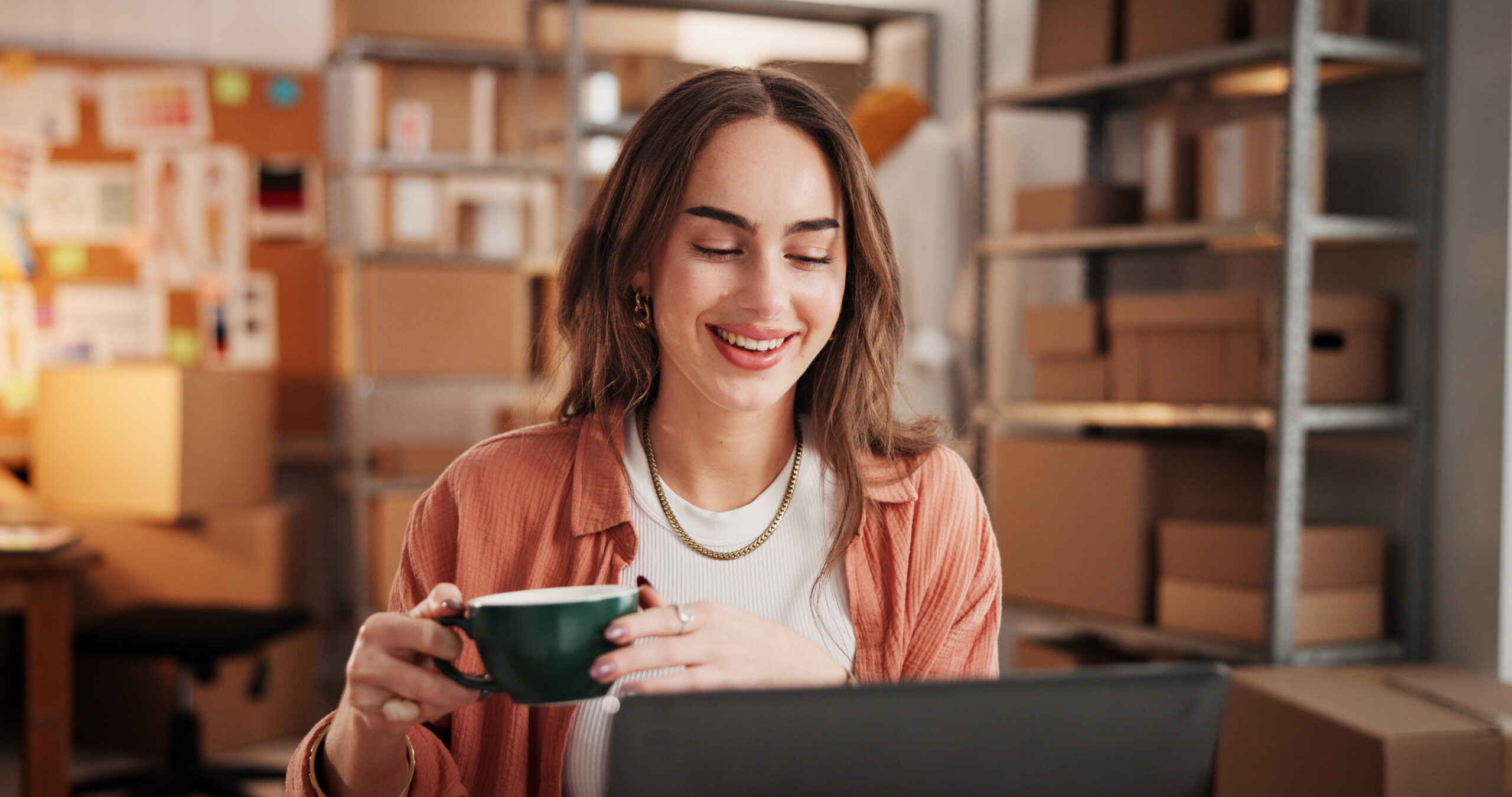 Woman, coffee and smile with laptop at small business for ecommerce, supply chain and online order. Female person, morning beverage and pc for logistics, inventory management and schedule shipping